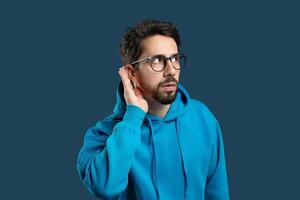 A man with a beard and glasses, dressed in a blue hoodie, stands against a solid color background. He appears focused as he cups his hand behind his ear, trying to hear something more clearly. photo