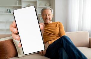 An older man relaxes on a couch in a well-lit room, smiling at the camera while holding up a blank smartphone. The setting suggests comfort and modern technology. photo