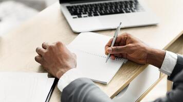 A person's hand writes in a notebook while sitting at a desk, a laptop in the background. photo