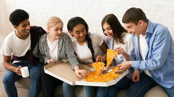 A diverse group of teenagers is sitting together, smiling and enjoying a large pizza. They are casually dressed, indicating a relaxed and social atmosphere photo