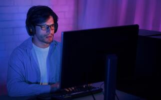 A young man with headphones on is sitting in front of a computer screen in a dimly lit room. He is wearing glasses and appears to be focused on the screen. photo