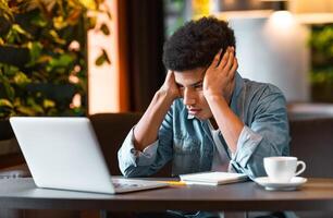 A young black guy is sitting at a table in a cozy cafe, looking frustrated as he works on his laptop. He is holding his head with both hands and has a notebook and a cup of coffee nearby. photo