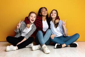 Three young multiethnic girls sit closely side by side with their backs against a bright yellow wall. They are casually dressed in jeans and flannel shirts and are smiling joyfully photo
