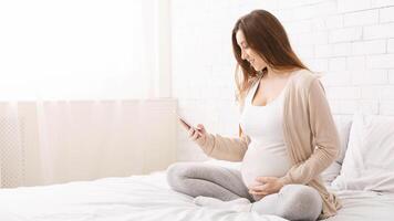 Cheerful pregnant woman browsing on smartphone, sitting on bed at home, empty space photo
