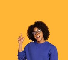 A young man with glasses and an curly hairstyle is smiling and pointing upward with his right hand. He is wearing a blue sweater. The background is a solid yellow color, copy space photo