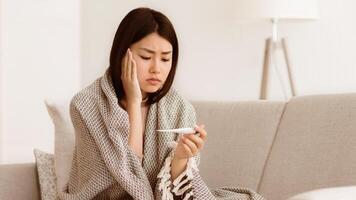 A woman wrapped in a blanket sits on a couch and checks her temperature with a thermometer. She looks concerned. photo