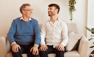 Joyful Elderly Father And Mature Son Smiling Each Other Sitting On Couch At Home. Selective Focus photo