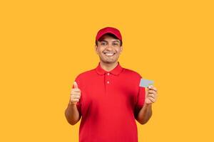 A cheerful delivery man dressed in a red uniform and cap gives a thumbs up while holding a card, standing against a bright yellow background. photo