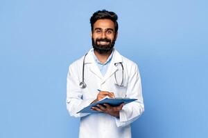 A smiling Indian man doctor with a beard is standing against a blue background. He is wearing a white lab coat and a stethoscope around his neck. He is holding a clipboard photo