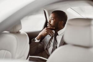 African American businessman in a suit and tie sits in the backseat of a car, looking out the window. He is deep in thought, with his hand on his chin. photo
