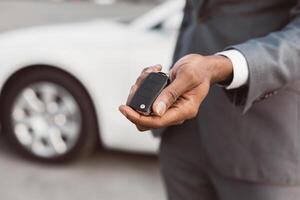 Black mans hand holding a car key in front of a white sedan. The man is wearing a gray suit and the key is a black remote. The car is partially out of focus and the mans hand is the main focus. photo