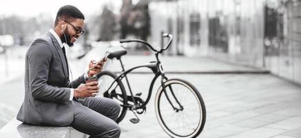 An African American man is using his phone while sitting next to his bicycle. The setting is outdoors, providing a blend of work and leisure in a cityscape, web-banner photo