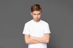 A teenager in a white t-shirt exudes a stern demeanor with arms crossed, displaying a no-nonsense attitude, isolated on a grey background photo