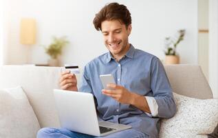 A man sits on a couch, smiling while looking at his smartphone. He holds a credit card in his other hand, and a laptop rests on his lap. photo