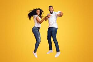 Smiling African American Couple Is Taking Selfie While Jumping, pointing at camera, yellow studio background photo