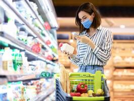 Young woman in disposable face mask taking dairy products from shelf in the supermarket, holding bottle and smartphone, scanning bar code on product through mobile phone, walking with trolley cart photo