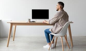 Rear Back View Of Happy Smiling Arab Man Looking At Pc Computer With Blank Black Screen For Mock Up Template Sitting At Desk At Home Office, Free Copy Space. People, Technology, Remote Work Concept photo