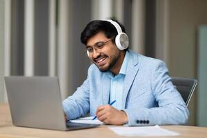 A smiling Indian man wearing headphones sits at a desk and works on a laptop computer. He is taking notes with a pen on a notepad and smiling. There are papers and a pen on the desk. photo