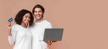 Easy Payments Concept. Happy multiracial spouses holding laptop and credit card, posing together over yellow studio background, copy space photo