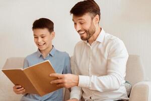 Happy Father And Son Reading Book And Laughing Sitting Together On Couch At Home. Parenthood And Family Relationship. Selective Focus photo