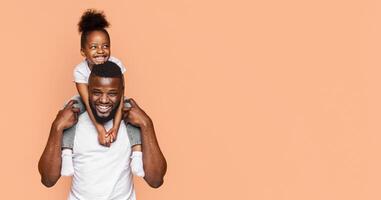 A happy black father and daughter pose for a picture against a peach-colored background. The little girl is sitting on her fathers shoulders, and both are smiling brightly at the camera, copy space photo