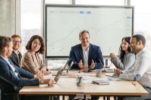 A team of five business professionals sit around a large wooden table in a modern office. The man at the head of the table is presenting a large graph on the screen behind him photo