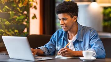 A young African American guy is seated at a table in a cafe, focused on his laptop screen. He is typing and clicking on the keyboard, surrounded by cups of coffee and notebooks. photo