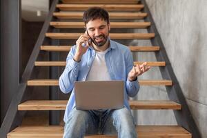 A man is sitting on wooden stairs in a home, talking on a cell phone while holding a laptop on his lap. He is gesticulating with one hand as he speaks, likely discussing something related to his work. photo