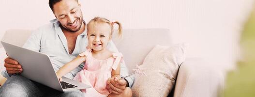 Family Weekend Concept. Little girl with wand sitting with father on couch using laptop. Empty space photo