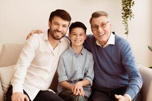 Male Generation. Happy Grandfather With Son And Grandson Smiling At Camera Hugging Sitting Together On Couch At Home. photo