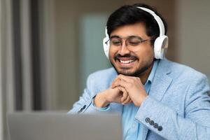 Indian man is smiling while wearing headphones and looking at a laptop screen. He is seated indoors with natural light coming in through a window to the left of the frame. photo