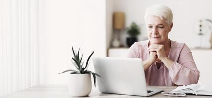 Old interested woman reading article online, using laptop at home, copy space photo