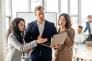 Three business professionals stand in a modern office, gathered around a laptop. They are all attentively listening and appear to be engaged in a productive conversation. photo