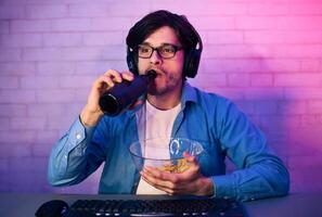 A young man wearing headphones and glasses is sitting at a desk, drinking from a black bottle, and eating chips from a bowl. He is likely playing a game as the desk has a keyboard and mouse. photo