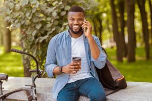 African American Man Employee Drinking Coffee In Park And Talking On Cellphone, Relaxing Outdoors During Break At Work photo