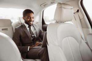 African American man in a business suit is seated in the back seat of a car, working on a laptop. He is smiling and appears to be focused on his work photo