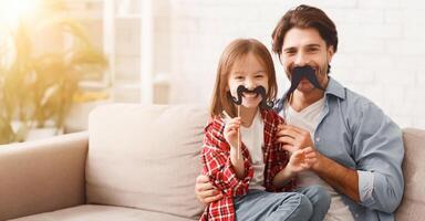 Beautiful funny little girl and daddy have mustaches on sticks, posing at home. Family holidays and togetherness concept, copy space photo