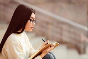 Asian woman is seated, focusing intently on writing in a notebook. Her hand is moving across the page as she concentrates on the task at hand, copy space photo