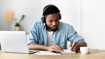Concentrated african american guy in headphones studying foreign language online through conference and making notes in notebook, panorama with copy space photo