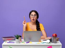 Idea Concept. Excited Young Indian Woman In Eyeglasses Sitting At The Desk Using Laptop And Raising Pencil Up Isolated Over Purple Background In Studio. Amazed Shocked Lady Having Insight, Aha Moment. photo