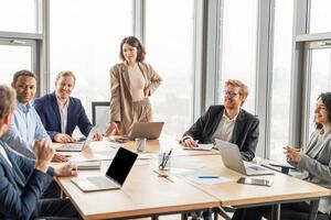 A diverse team of business professionals are gathered around a conference table in a modern office, engaging in a lively discussion. The woman standing at the head of the table, leading the meeting photo