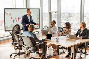 A businessman is presenting financial data to a team in a modern office with large windows overlooking the city. He is standing and pointing at a chart on a screen photo