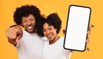 A young black couple with big smiles stand in front of a yellow background, holding a smartphone with a blank screen. The image is perfect for displaying a mobile app, website, or product. photo