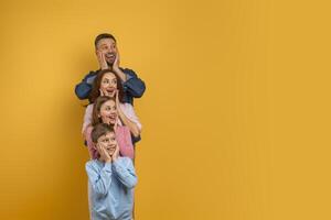 Surprised family of four, with two children and their parents, is lined up vertically, each placing their hands on their cheeks in a playful expression of surprise and happiness, copy space photo
