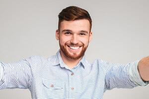 Cheerful handsome bearded redhaired man holding camera and making selfie on beige background, closeup photo