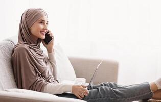 Smiling Muslim Girl In Hijab Having Phone Conversation And Using Laptop While Sitting On Sofa Against Window At Home, Free Space photo