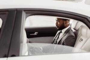 Black man in a suit is sitting in the back seat of a white car. He is looking down and appears to be thinking or working on something. The car is parked in a bright location, likely outdoors. photo