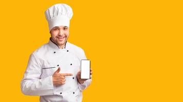 Smiling Chef Man Showing Smartphone Empty Screen Recommending Food Delivery Mobile App Standing Over Yellow Studio Background. Mockup, Panorama photo