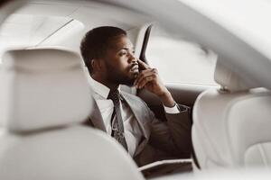 An African American businessman sits in the back seat of a car, wearing a suit and tie. He is looking out the window, lost in thought. He has a hand on his chin photo