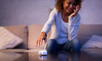 Depression. Crying afro woman taking antidepressant pills, sitting on sofa photo